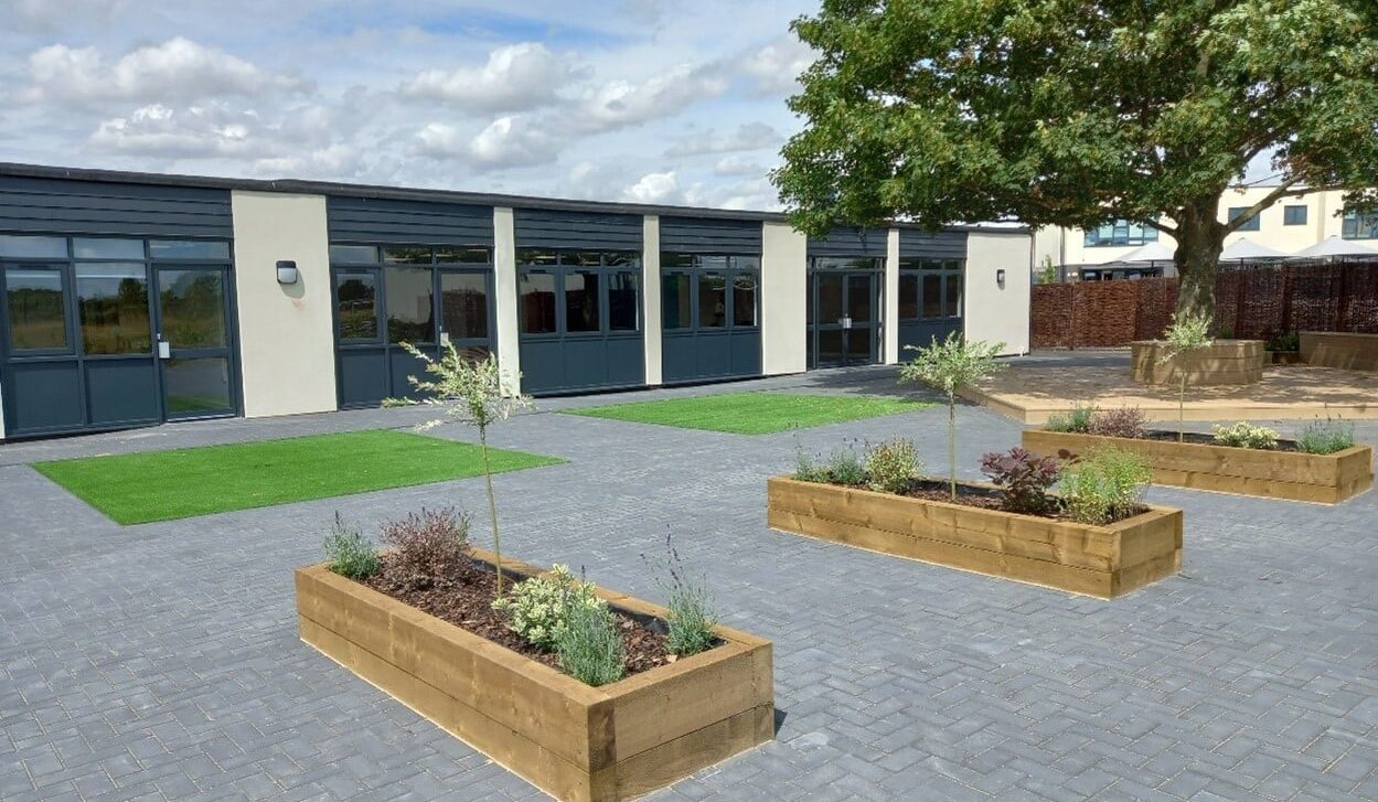 Single storey, modern school building with big, dark windows facing a paved area and planters